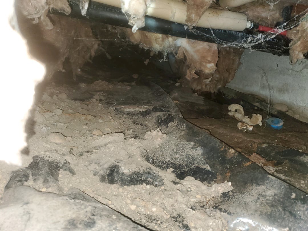 Crawl space with exposed soil, an old ground liner and hanging insulation around pipes